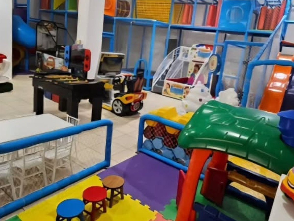 Área de brinquedos com estrutura de escalada azul, escorregador, piscina de bolinhas, mesa de air hockey e videogame