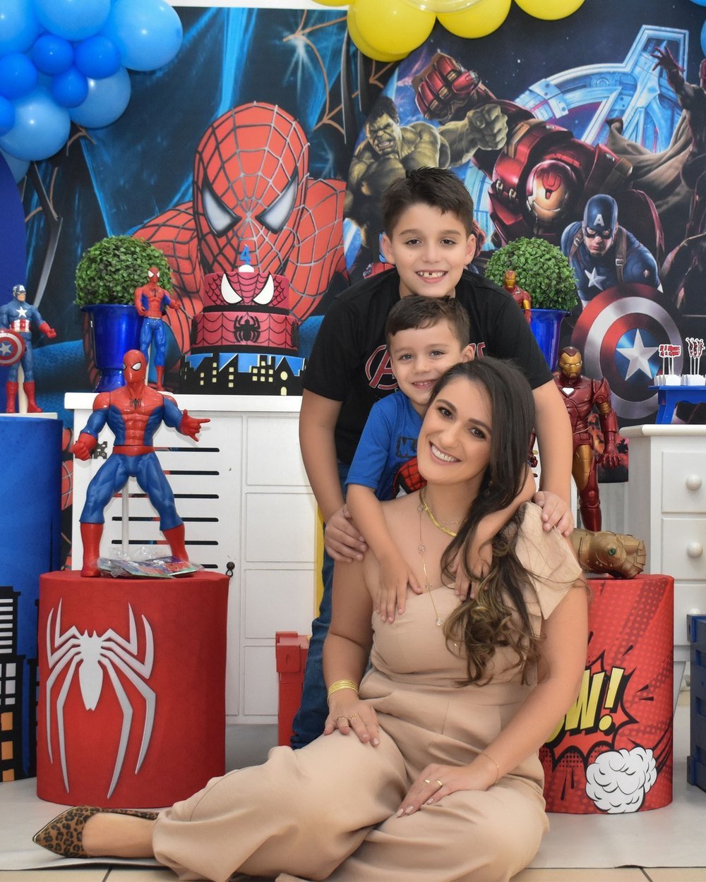 Mãe sentada no chão sorrindo, abraçada pelos dois filhos, na frente da decoração de festa montada