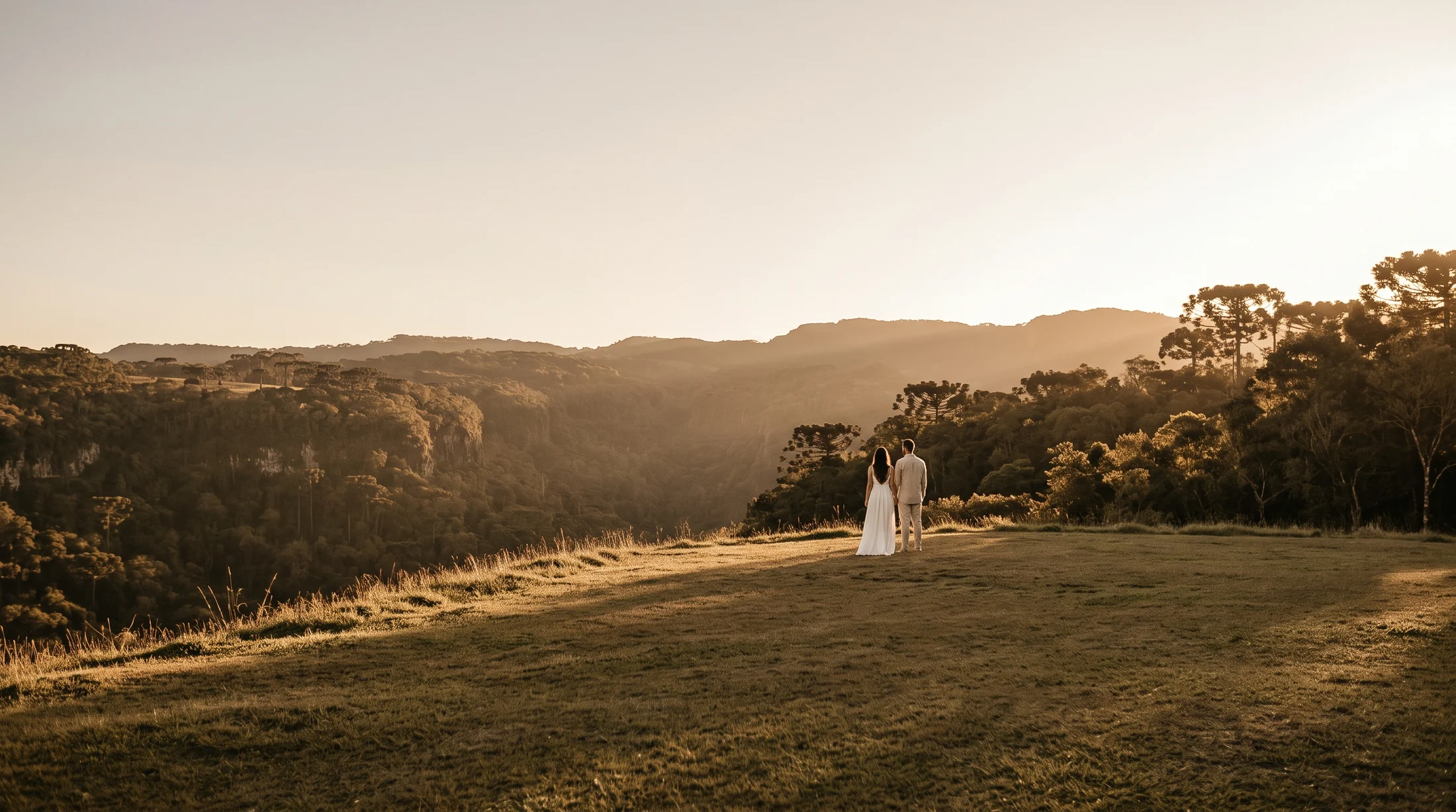 Casal ao longe num gramado aberto, diante do vale da serra na hora dourada