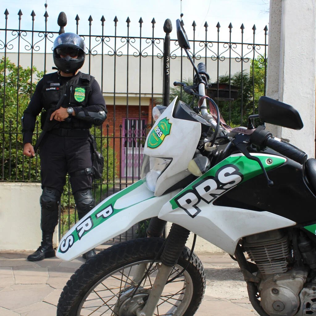 Agente da PRS fardado ao lado da moto de ronda em frente a um portão
